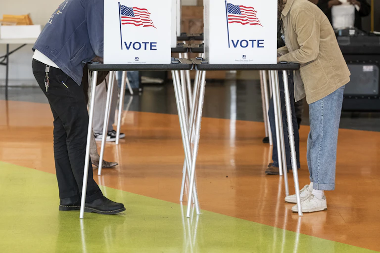 Two casually dressed voters lean into opposite voting stands with their faces obscured as they complete ballots