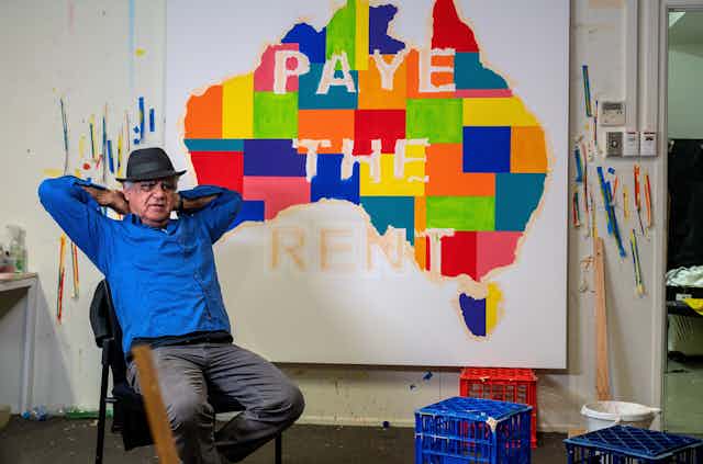 Richard Bell in front of a painting reading 'pay the rent'