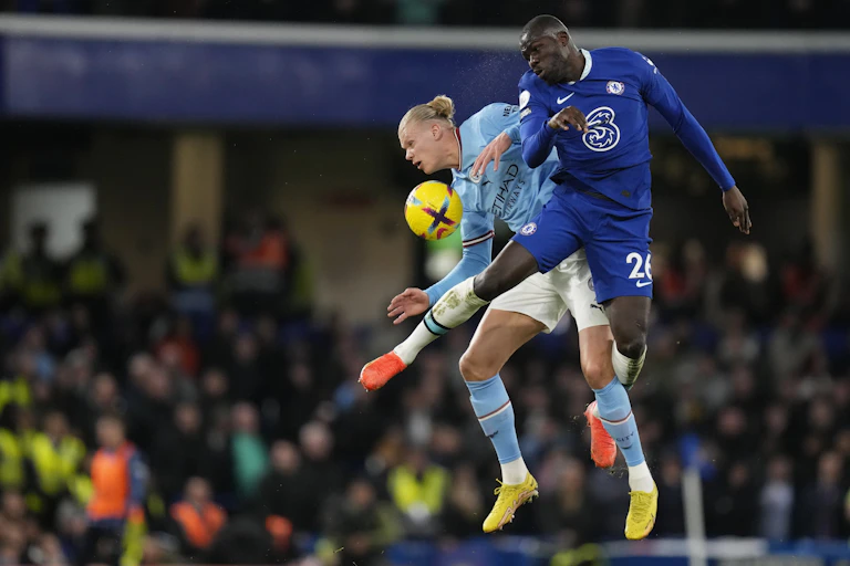 Two men - one in a light blue uniform and one in dark blue - captured in a jump mid-air with a yellow ball in the foreground