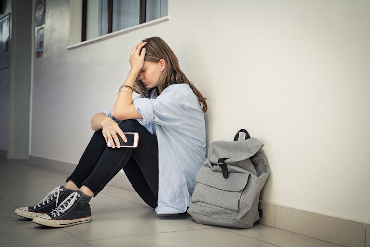 A sad or stressed young woman sits on a hallway floor holding her head.