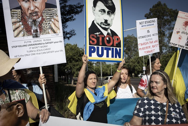 Women protestors hold up signs, one saying 'Stop Putin' with a picture of Putin as Hitler.
