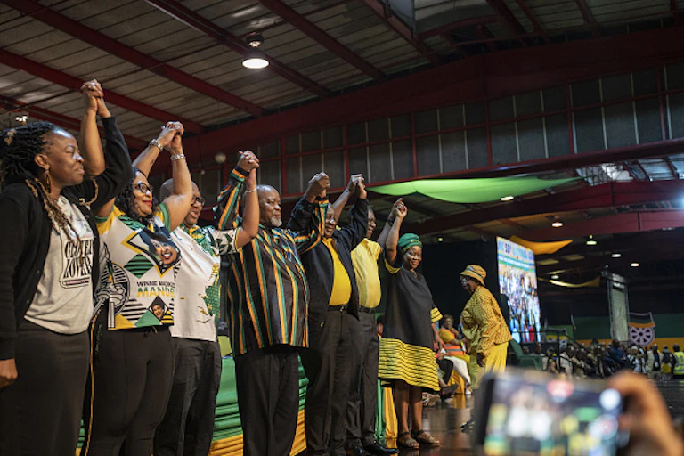 Three women and four men standing on a stage and raising interlinked hands in greeting at a meeting..