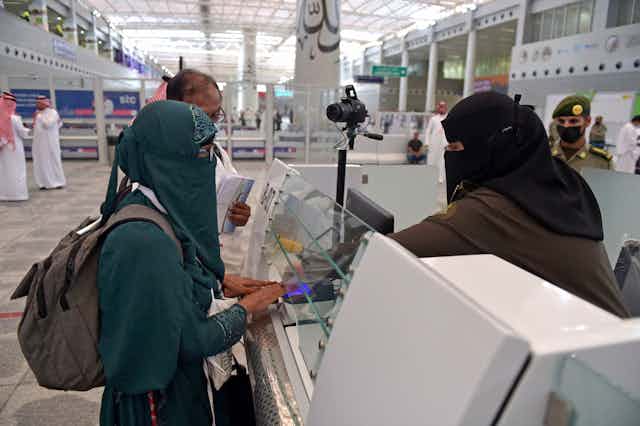 A veiled woman presents her passport to a veiled screener at a counter.