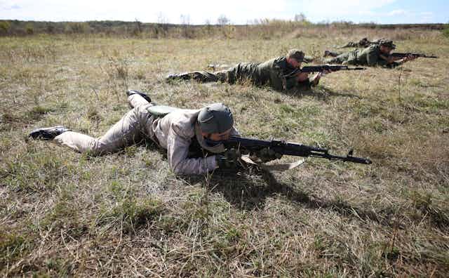 Men in combat gear carrying weapons inch along the ground on their stomachs in a training exercise.