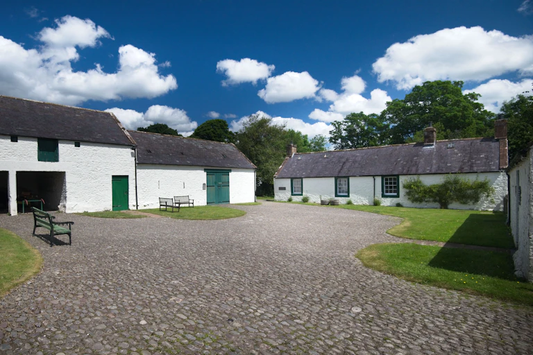 A cobbled farm courtyard surrounded by whitewashed byres and sheds against a bright blue sky with white clouds.