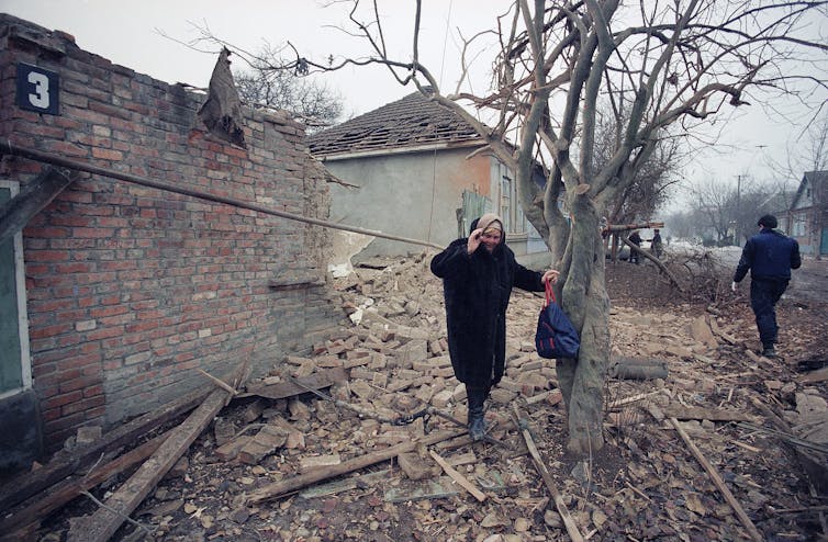 An elderly woman holds onto a tree as she looks at the rubble surrounding her.