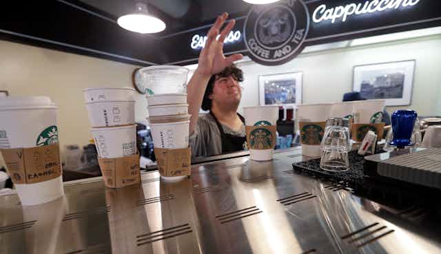 Several white paper cups with paper holders sit atop a coffee machine with a barista holding his right hand up in the background inside a coffee shop