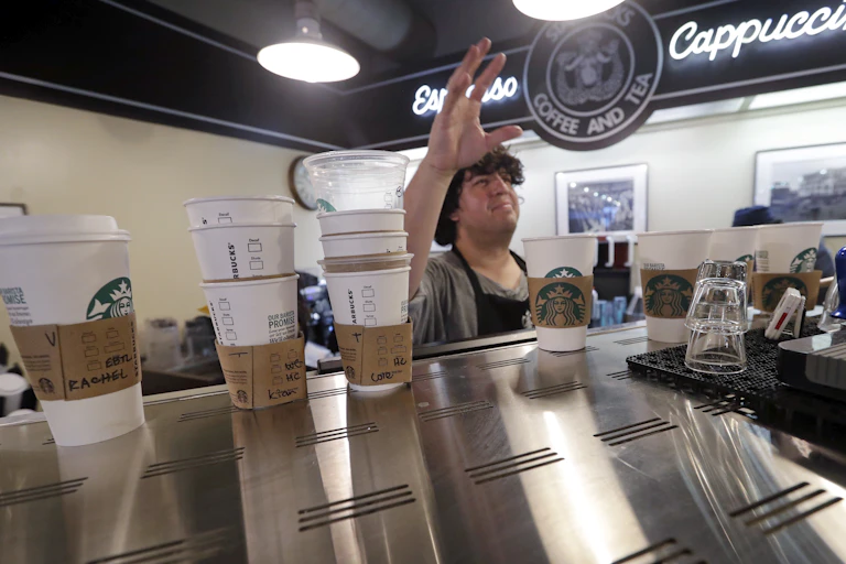 Several white paper cups with paper holders sit atop a coffee machine with a barista holding his right hand up in the background inside a coffee shop