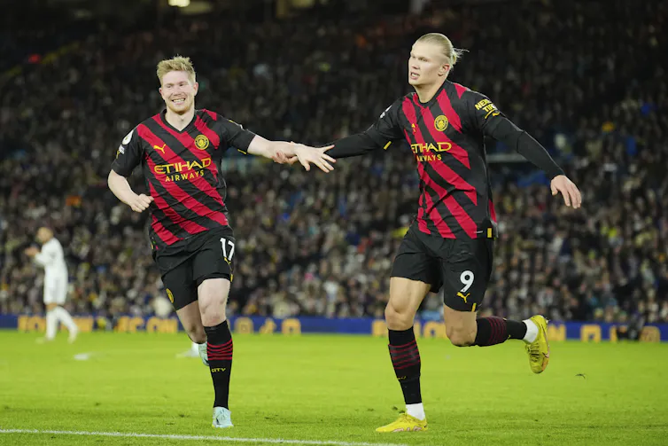 Two soccer players wearing black and red outfits running on a pitch.