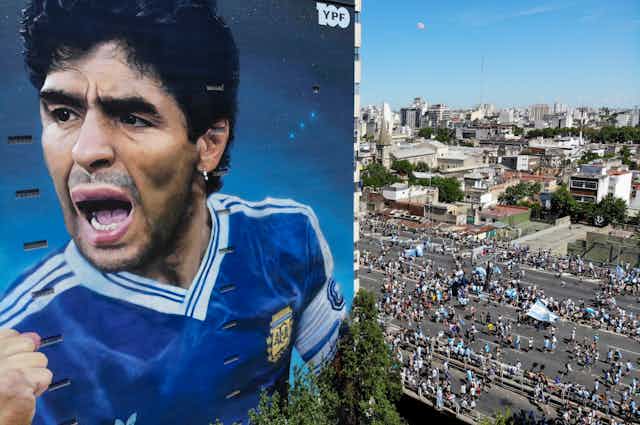 A large mural of Diego Maradona on the side of a building. People carrying Argentine flags walk past.