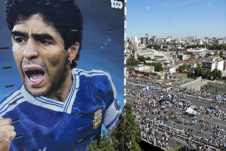 A large mural of Diego Maradona on the side of a building. People carrying Argentine flags walk past.