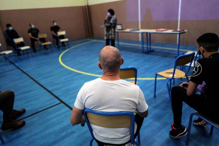 A few men sit at classroom desks in a simple room with a blue floor.