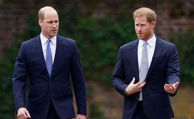 Two men in navy suits, one bald and one with red hair, speak as they walk beside each other.