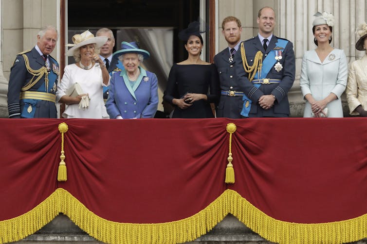 Various Royal Family members stand on a balcony festooned with red and gold bunting looking out at the crowds. An elderly woman dressed in blue is in the centre.