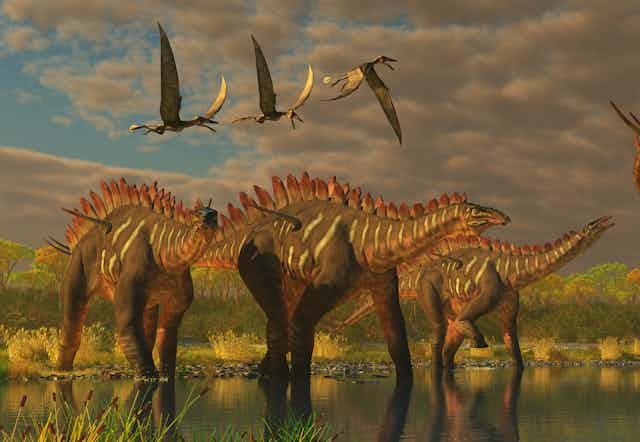 Flying dinosaurs are in the air over other dinosaurs standing in marshland