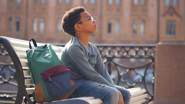 A boy sits on a bench with his school bag