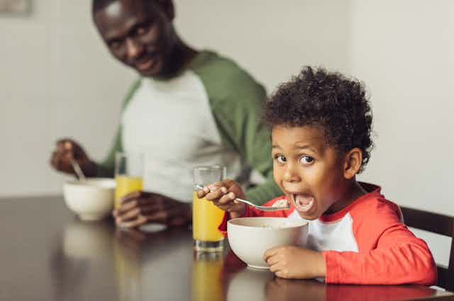 young boy is eating cereal from a spoon with father looks on