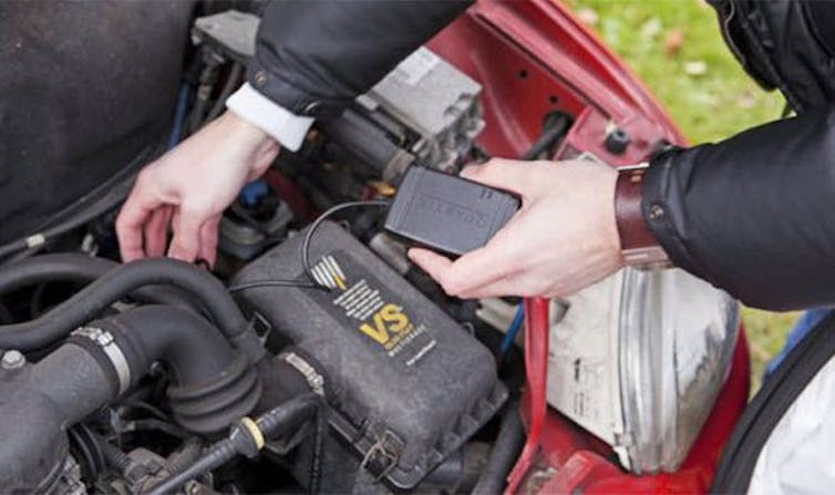 A pair of hands, one holding a black box near a car engine