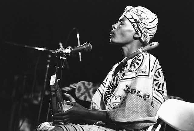A black and white photo of a woman on stage. She wears a head scarf and traditional African attire, her eyes closed as she plays on a thumb piano, a microphone positioned near her mouth.