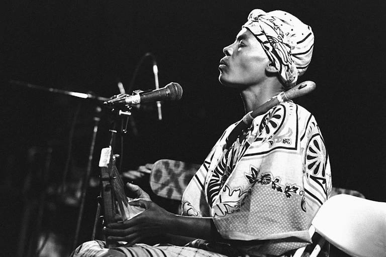 A black and white photo of a woman on stage. She wears a head scarf and traditional African attire, her eyes closed as she plays on a thumb piano, a microphone positioned near her mouth.