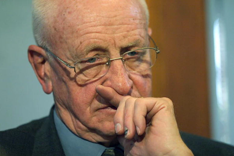 A balding old man wearing specs is in deep thought, with his left partly covering his mouth.