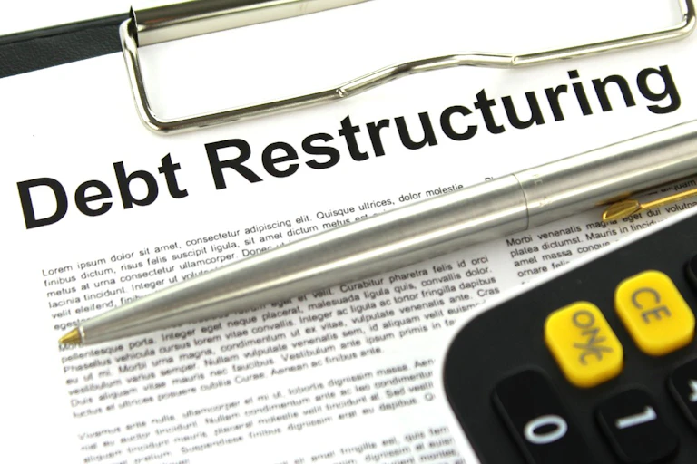 A piece of printed paper on a clipboard with debt restructuring as the headline. A silver pen and a section of a black calculator lie on top of the paper.