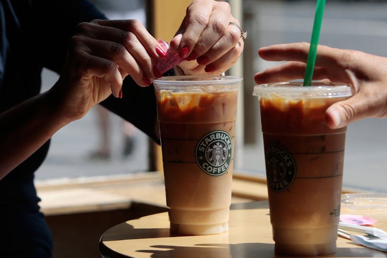 Several hands hold two clear plastic cups of iced coffee on a table