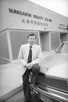 A man in dark pants and a light blazer sits on a car in front of a building with 'Mandarin Realty Co. Inc' written on it.