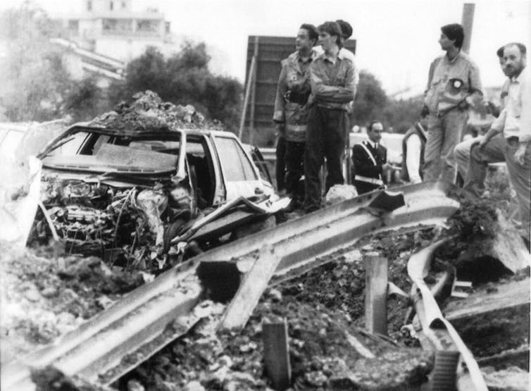 Men stand around the wreckage of a car following a bombing.