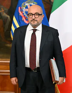Gennaro Sangiuliano stands in front of the Italian flag wearing a black suit and burgundy tie.
