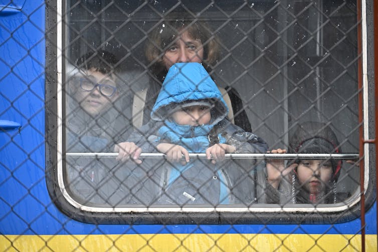 A blond middle-aged woman holds a child in a blue snowsuit up against her, next to two other older children, as seen through a window of a train and a large fence.