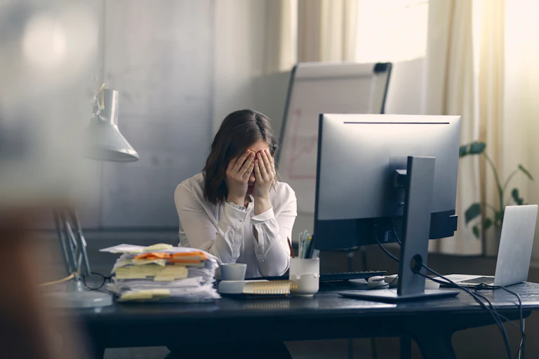 a woman sits in front of a computer screen with her head in her hands next to a pile of papers with sticky notes