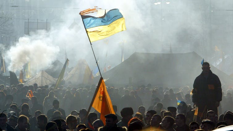 A person raises a blue and yellow striped flag above a large crowd of people, who are mostly covered in smoke.