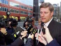 A dark haired man with blue eyes is at the centre of a media scrum.