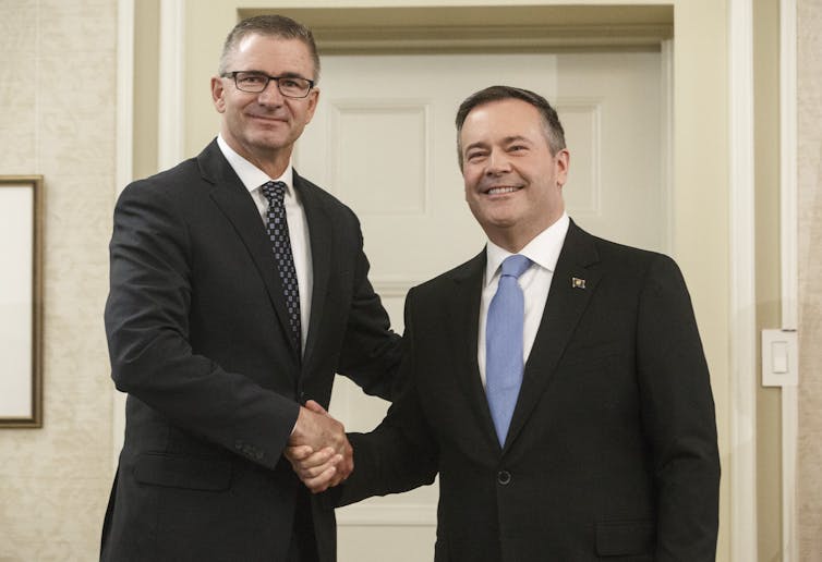 A tall man with glasses shakes hands with a shorter dark-haired man. Both are wearing suits and ties and smiling.