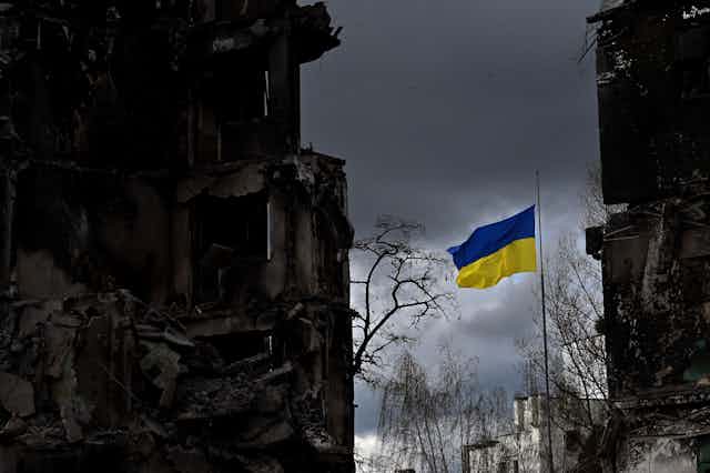 A blue and yellow flag waves against a dark grey sky, surrounded by dark buildings. 