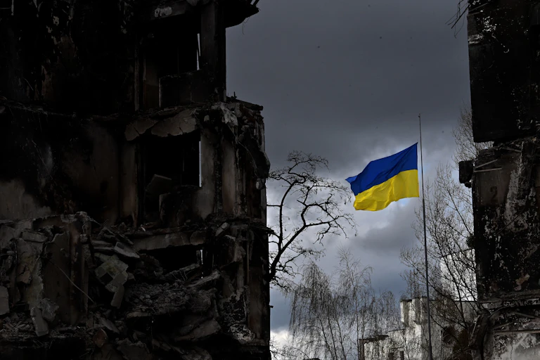 A blue and yellow flag waves against a dark grey sky, surrounded by dark buildings.