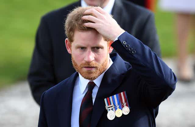 Prince Harry puts his hand on his head. He is wearing a dark blue suit and three military style medals.