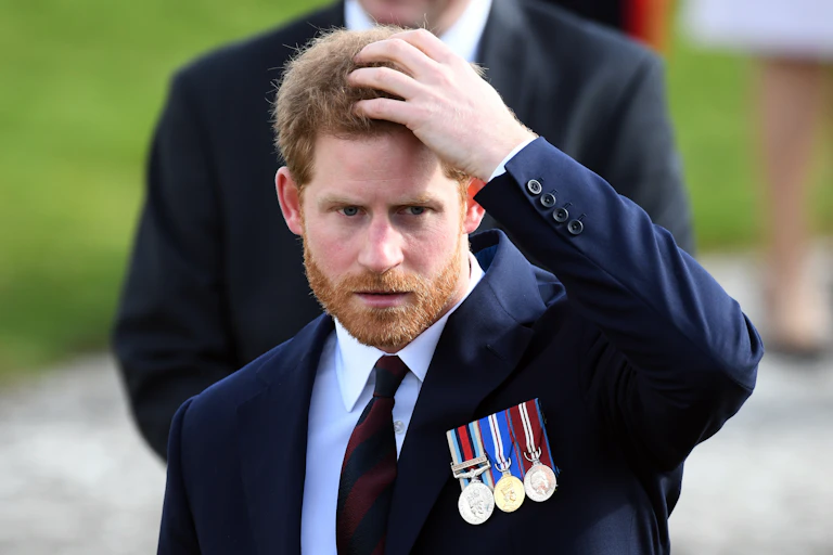 Prince Harry puts his hand on his head. He is wearing a dark blue suit and three military style medals.