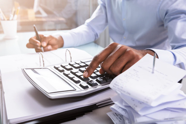 A person in a formal shirt, whose face is out of shot, uses a calculator and makes notes with a pen