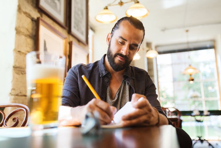 A man sitting in a cafe with a beer, writing notes.