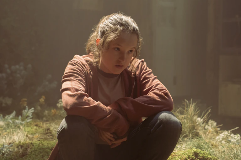 Bella Ramsey wears a pink hoodie and sits with her arms crossed.
