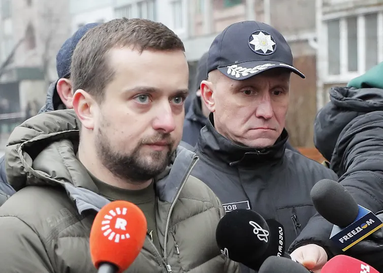 A beared man speaks into a journalist's microphone a man stands by wearing a baseball cap.