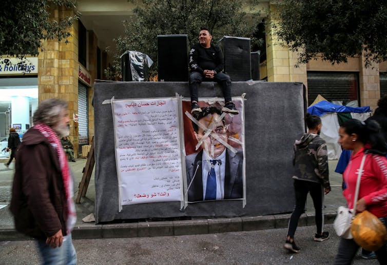 Un póster ilustrado desfigurado de un hombre en una caja grande, con un hombre sentado encima junto a los altavoces, mientras la gente pasa.