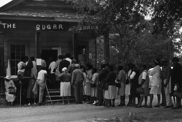 A group of about two dozen people lined up outside a rural store.