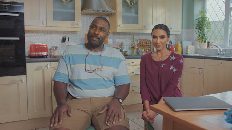 Deepfakes of Idris Elba and Kim Kardashian sit side by side in a kitchen in regular clothes.