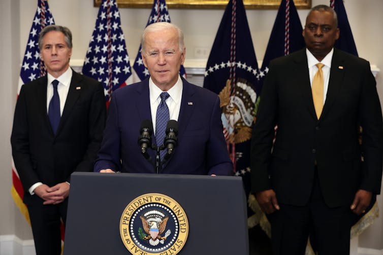 An older white man wearing a suit stands at a podium with the presidential seal on it . A middle aged black man and white man, both in suits, stand behind him, with American flags in the background.