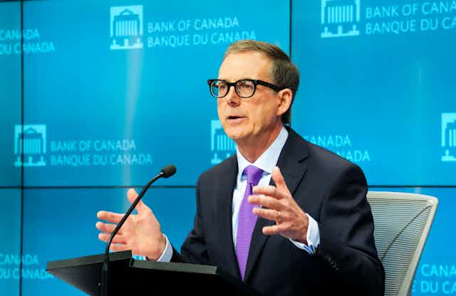 A man in a suit, purple tie, and black-rimmed glasses gestures as he speaks from behind a podium