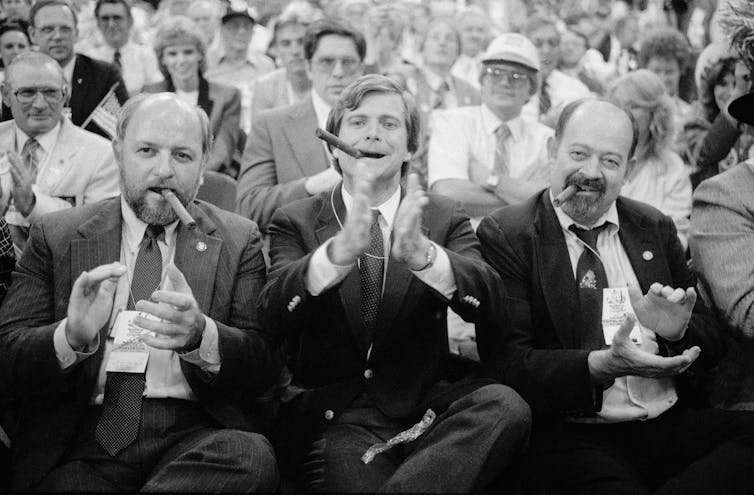 Three men in suits at a large gathering smoking cigars, clapping and looking happy.