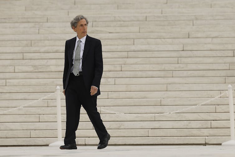 A gray-haired man in a suit walking in front of a lot of marble steps.